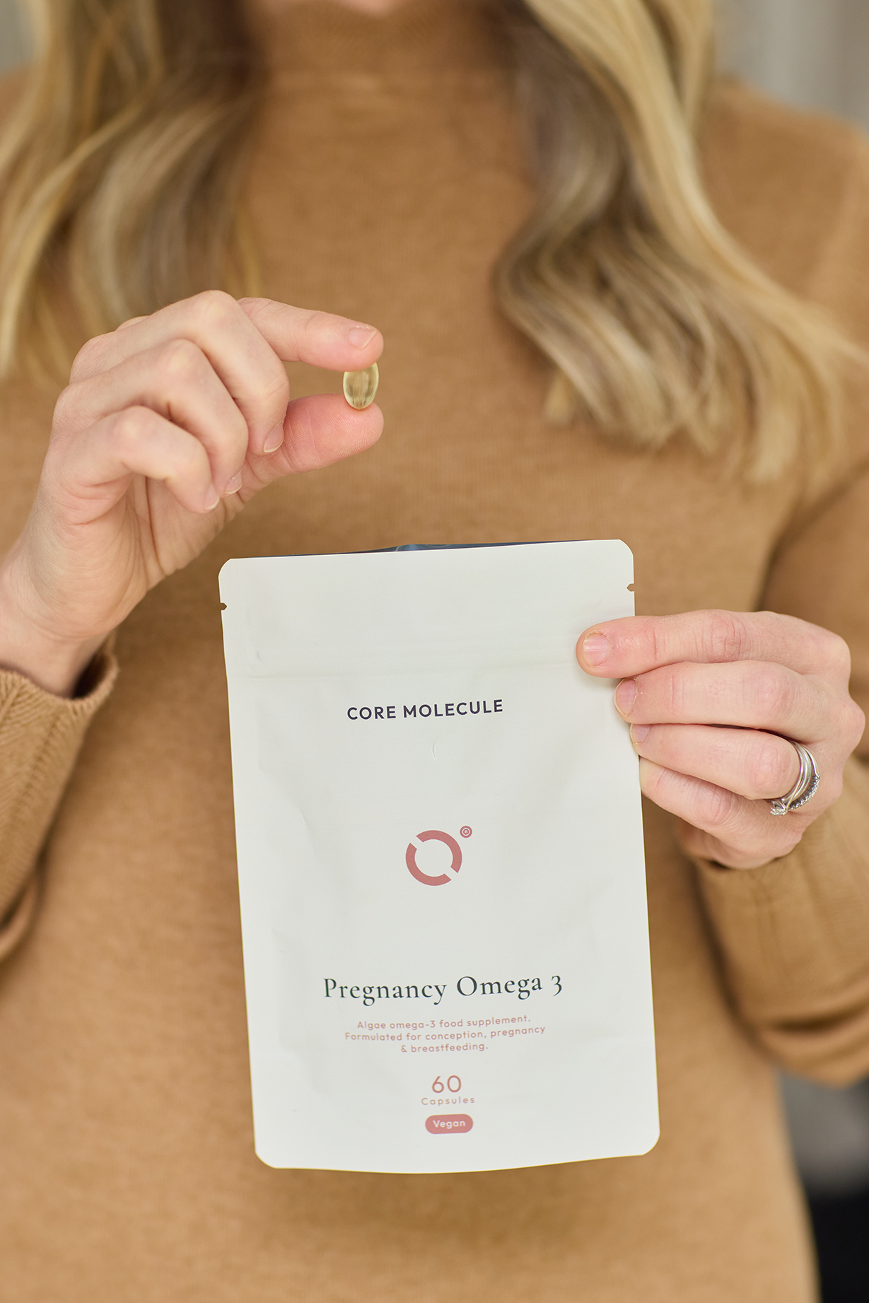 Woman holding a Core Molecule Algae Omega 3 tablet in her left hand and in the right hand she is holding thee 60 tablet recyclable packaging for the Core molecule Pregnancy food supplement. The woman is wearing a light brown jumper and you can just see her blond hair, but not her face.
